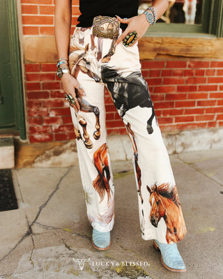 A person wearing wide-leg pants with a western horse print, styled with a black top, turquoise boots, and a silver belt, standing in front of a brick wall.