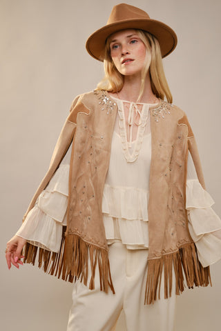 Woman wearing a beige fringe jacket, white blouse, and hat against a neutral background
