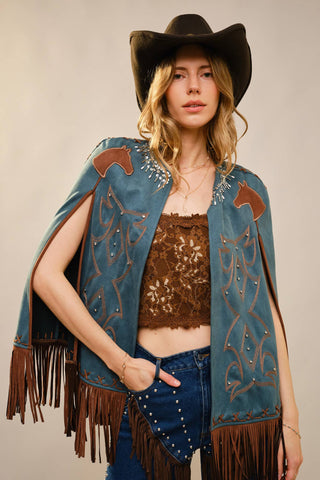 Woman wearing a cowboy hat, denim vest with fringe and embroidery, and brown top against a neutral background.