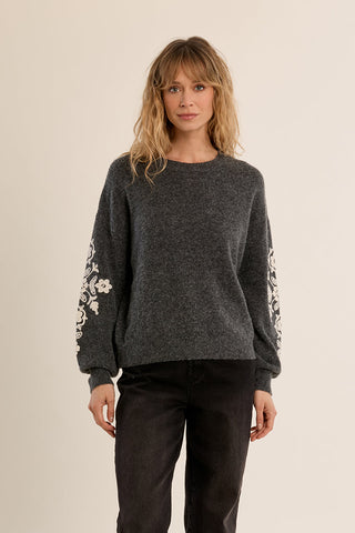 Woman wearing a dark grey knitted sweater with cream floral embroidery on the sleeves