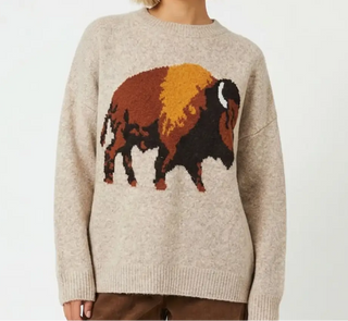 A person wearing a beige knit sweater with a large buffalo graphic across the chest in brown, orange, and black, paired with brown pants against a plain light background.