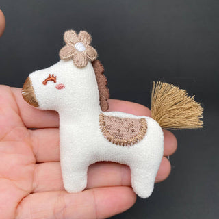 White fabric horse brooch with embroidered flower crown and golden fringe tail held in hand