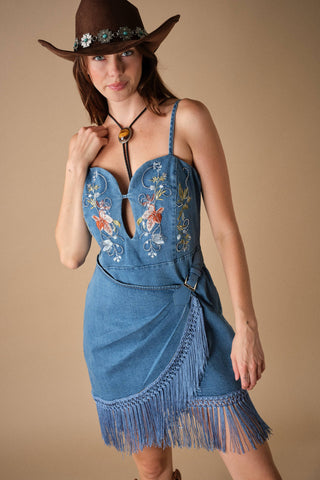 Model wearing embroidered denim mini dress with fringe hem, spaghetti straps, and wrap skirt