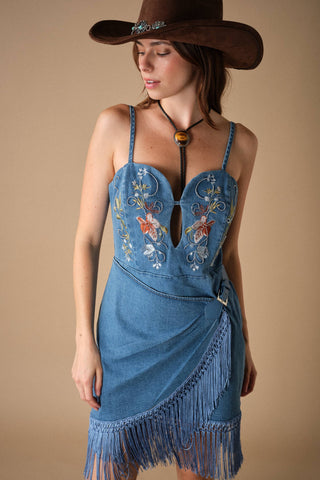 Model wearing embroidered denim mini dress with fringe hem, spaghetti straps, and wrap skirt