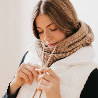 Woman wearing chunky knit tan cowl neck snood scarf with knitting needles