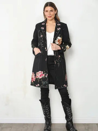 Woman wearing a black longline duster jacket with pink and white floral embroidery over a white top, black pants, and boots