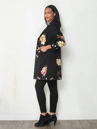 Side view of a woman wearing a black longline duster jacket with pink and gold rose embroidery, paired with black leggings and ankle boots