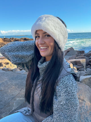 White faux fur headband ear warmer worn by woman at rocky ocean shore