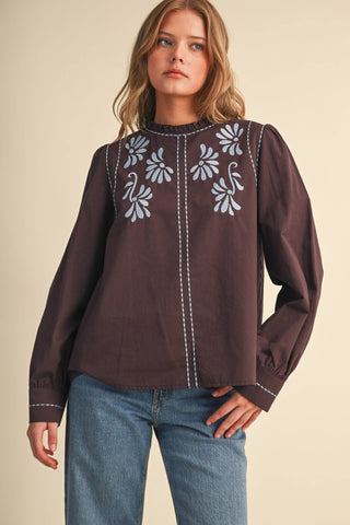 Woman wearing dark brown embroidered mock neck long sleeve top with blue floral details