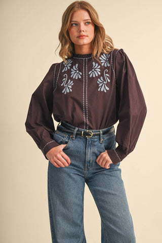 Model wearing dark embroidered mock neck top with puff sleeves and floral blue stitching detail
