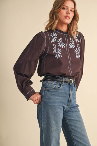 Model wearing dark brown embroidered mock neck blouse with floral motifs and long puff sleeves