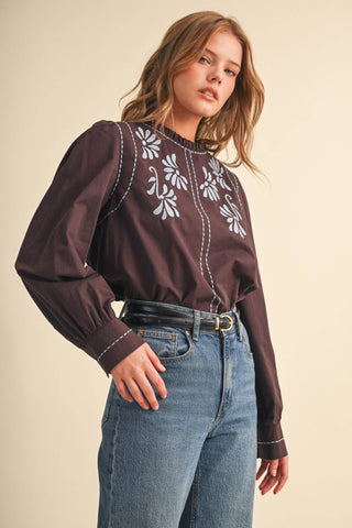 Model wearing dark brown long-sleeve mock neck blouse with white floral embroidery and jeans