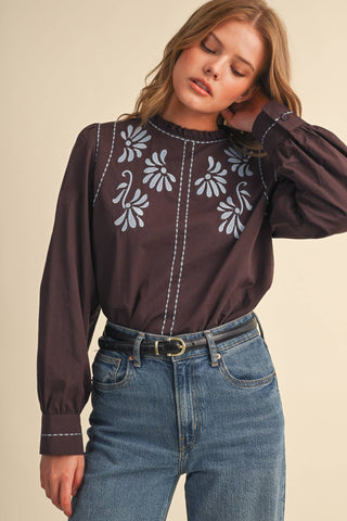 Model wearing dark brown embroidered mock neck blouse with floral motifs and long sleeves