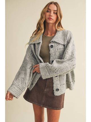 Model wearing an oversized grey eyelit knit cardigan sweater with button closure and wide collar