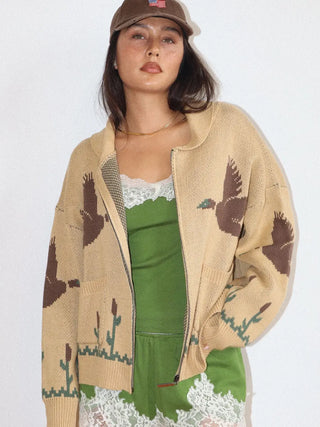 A woman models a beige knit zip-up hoodie featuring brown duck and green reeds pattern, worn over a green top and white lace skirt, with a brown cap.