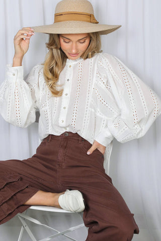 Model wearing white eyelet cotton blouse with dramatic puff sleeves and button front, seated pose